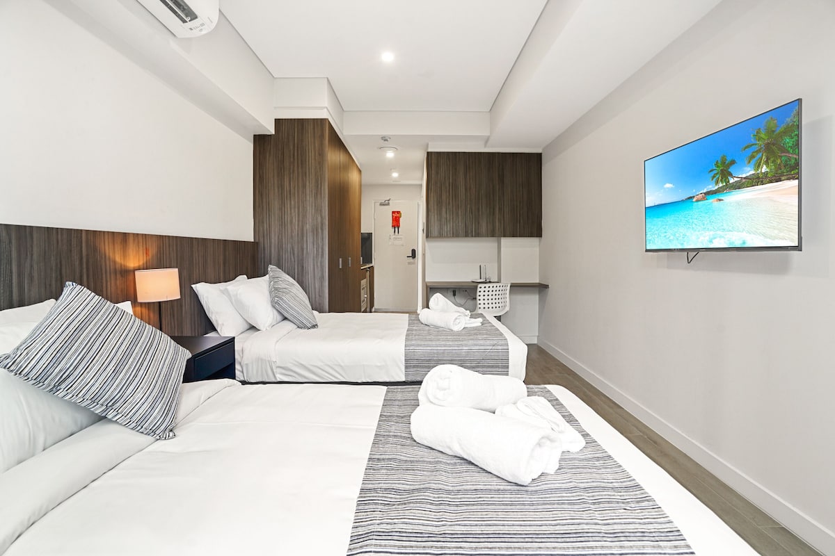 A hotel room with two beds, a TV, a bathroom, and a beach view.