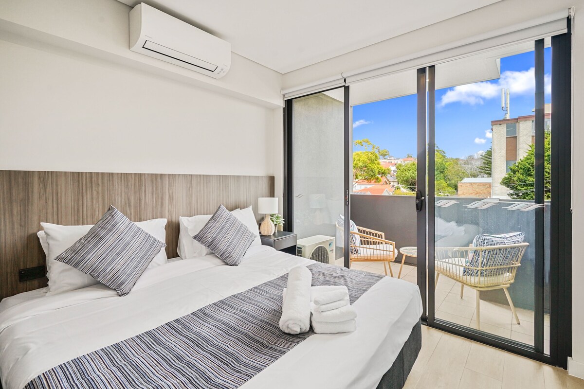 A modern hotel room with a large bed, a balcony with wicker chairs, and a city view.
