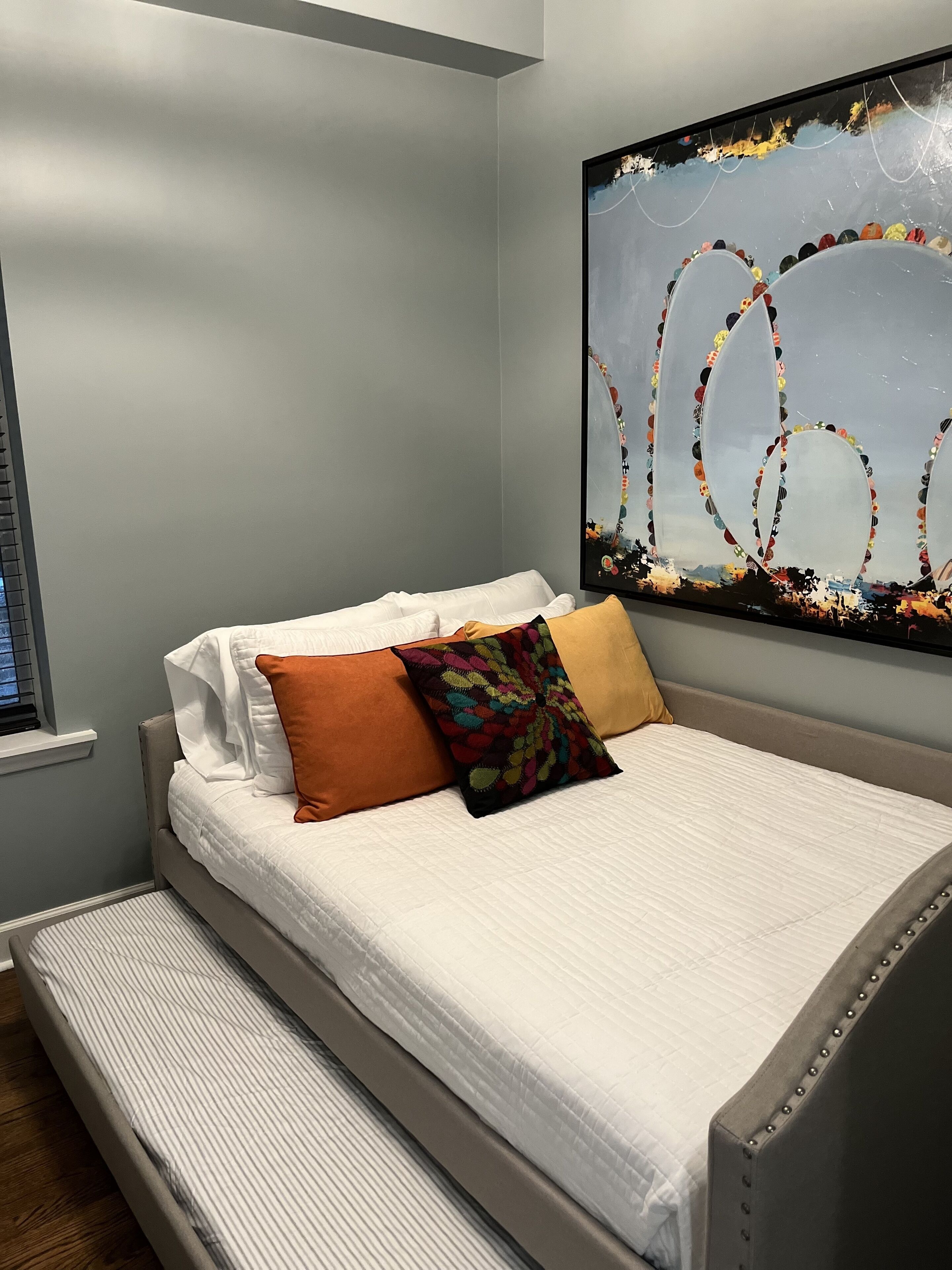 Una cama bien hecha con ropa blanca y dos almohadas, una marrón y otra multicolora, en una habitación con paredes grises y un cuadro abstracto grande sobre la cama.