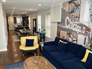 A modern living room with a blue sofa and yellow armchair, a dining area with a table and chairs, and a kitchen in the background.