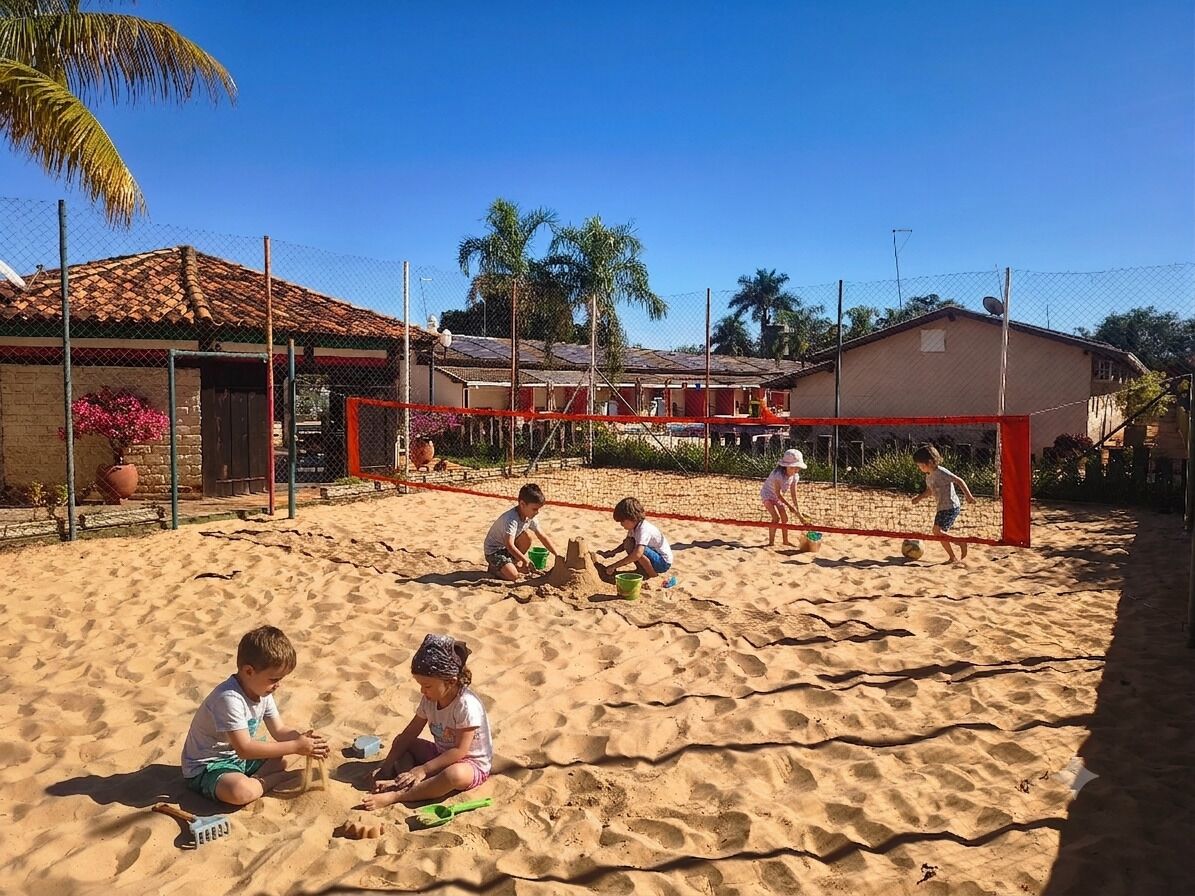 A sandy outdoor area with a volleyball net, surrounded by buildings and palm trees.