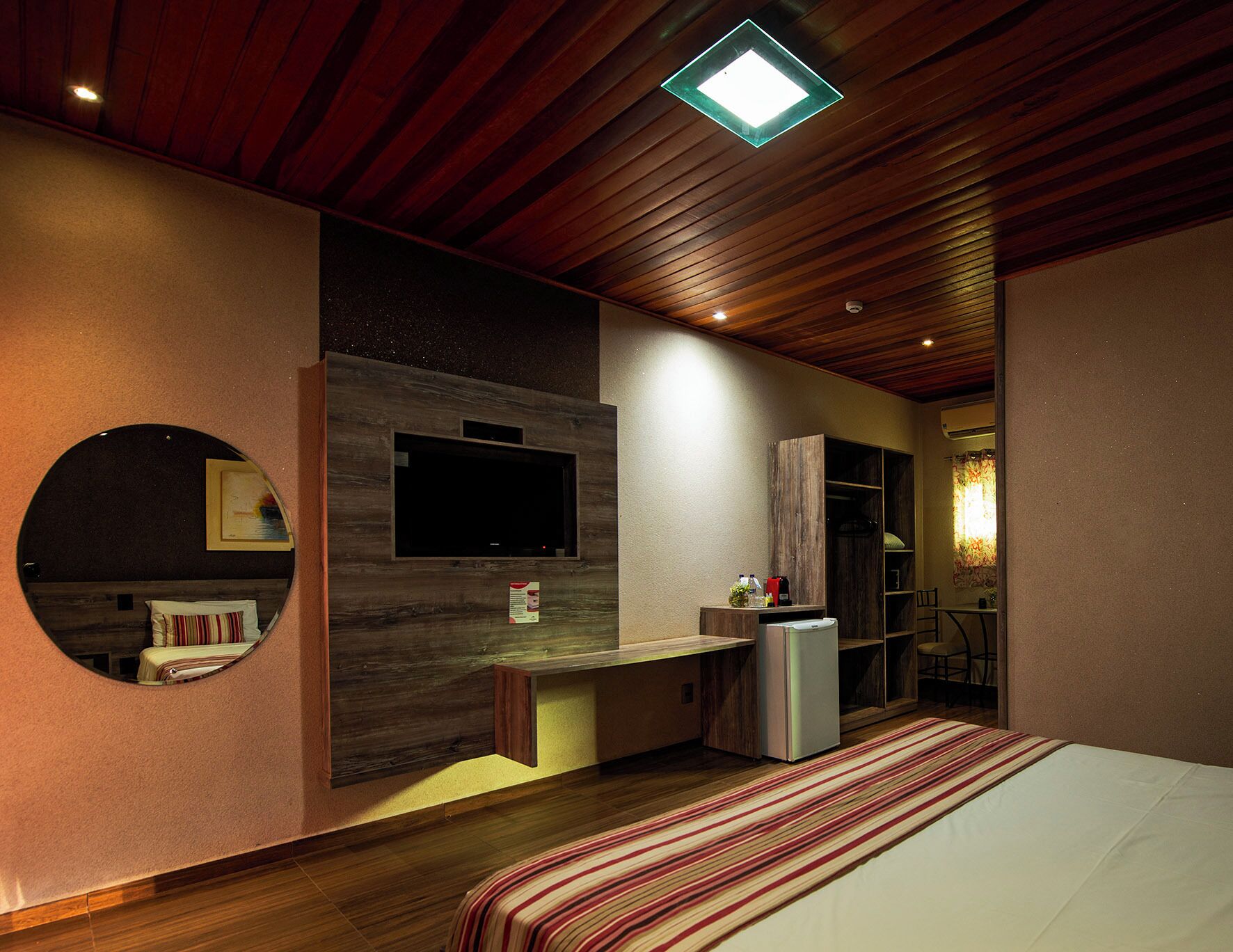 A modern bedroom with a large bed, a built-in TV, a small kitchenette, and a circular mirror.