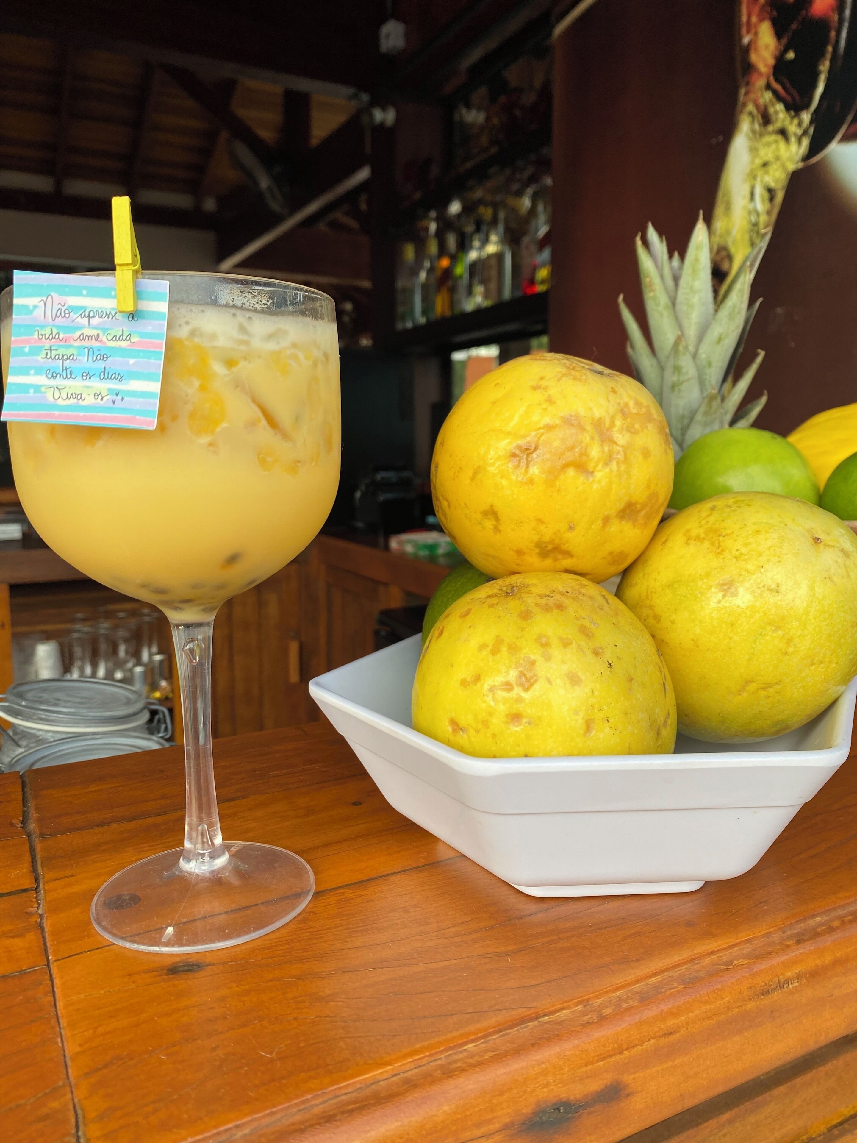 Een cocktailglas met een gele rietje en een briefje, een schaal met geel fruit, en een ananas op een houten tafel.