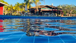 A swimming pool with clear water, surrounded by palm trees and a building with a tiled roof. - Hotel Pousada Villa Itália (Olímpia)