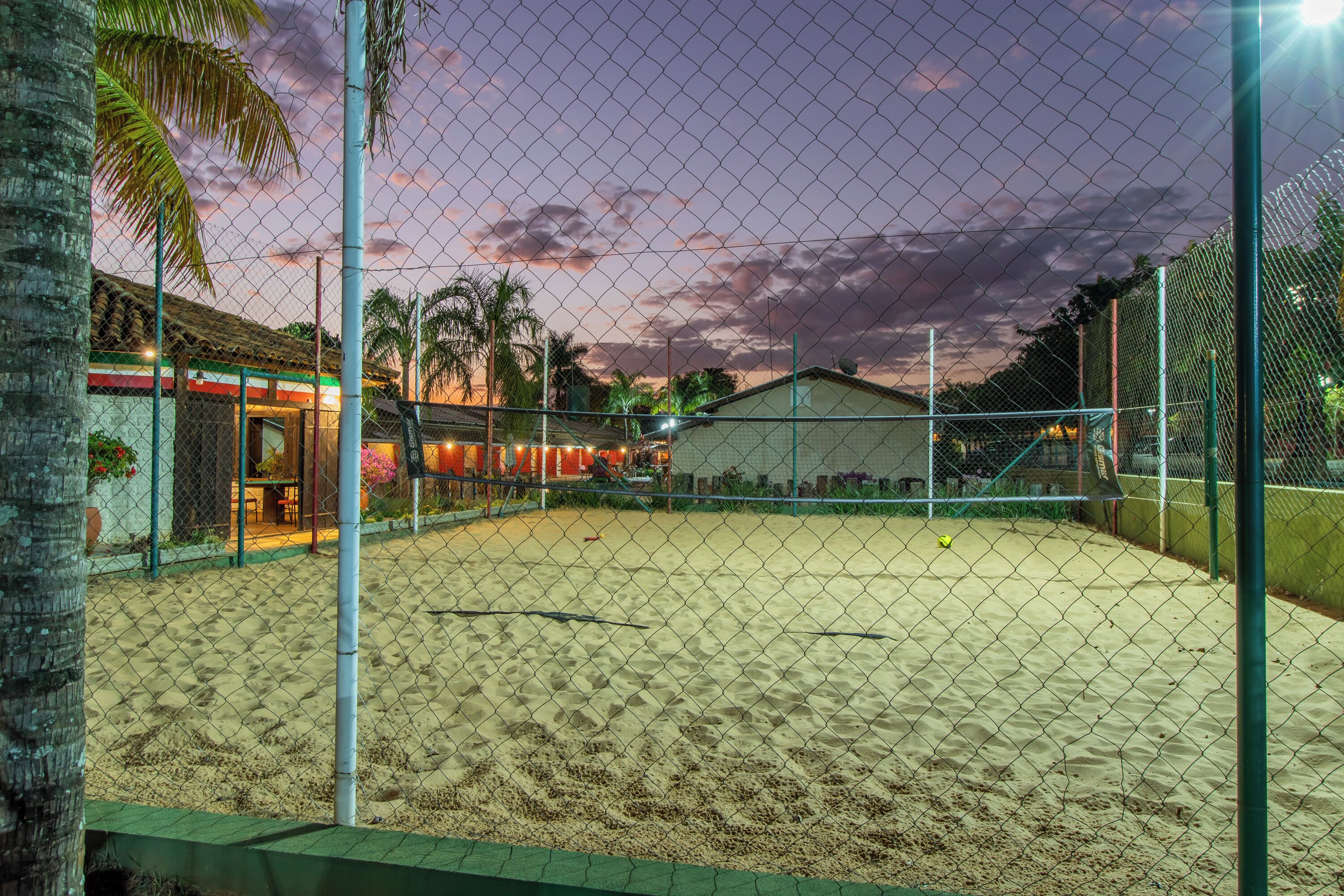 A fenced tennis court with a sandy surface, surrounded by a chain-link fence and palm trees.