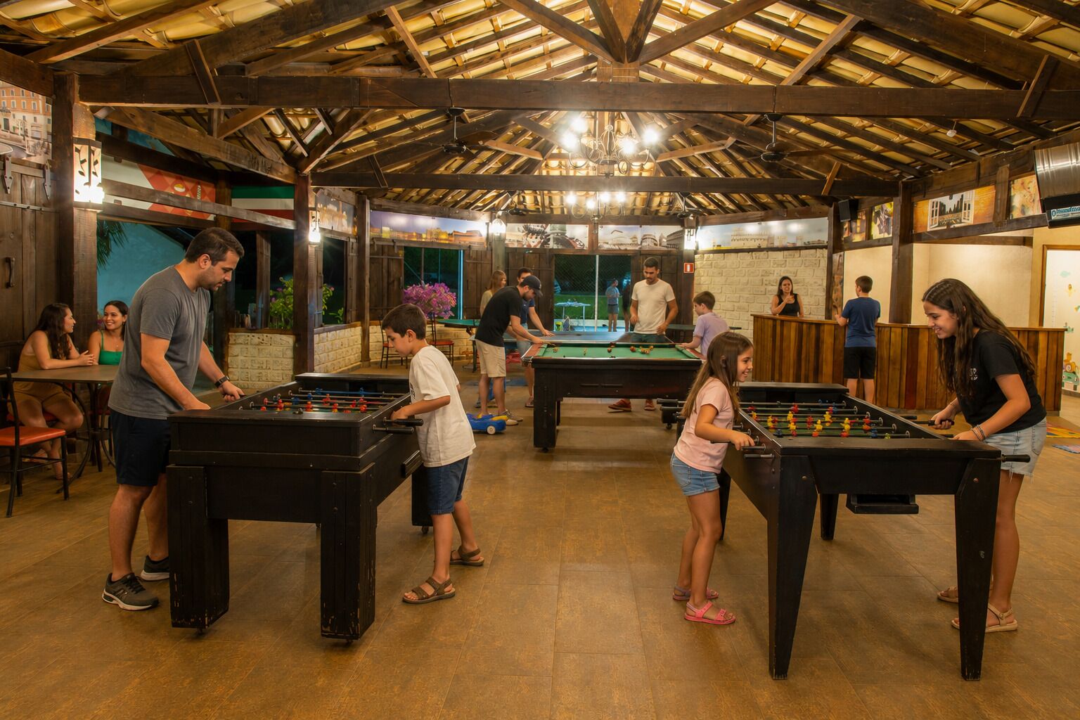 A recreational area with foosball tables and pool tables, wooden beams on the ceiling, and a brick wall.