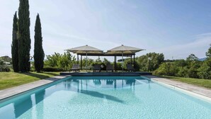 Ein rechteckiger Pool mit Holzterrasse, zwei hohe Zypressen und einem Pavillon mit Sonnenschirmen und Liegestühlen.