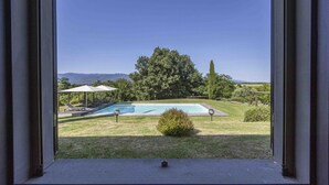 Blick durch ein Fenster auf einen Swimmingpool, eine Rasenfläche und ferne Berge.