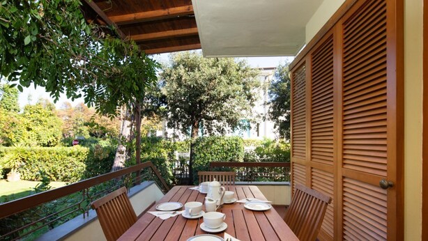 A wooden table set for four with plates, cups, and a pitcher, surrounded by chairs, on a balcony with a view of greenery and trees. - Villa Luia by MMega Villas  (Marina di Pietrasanta)