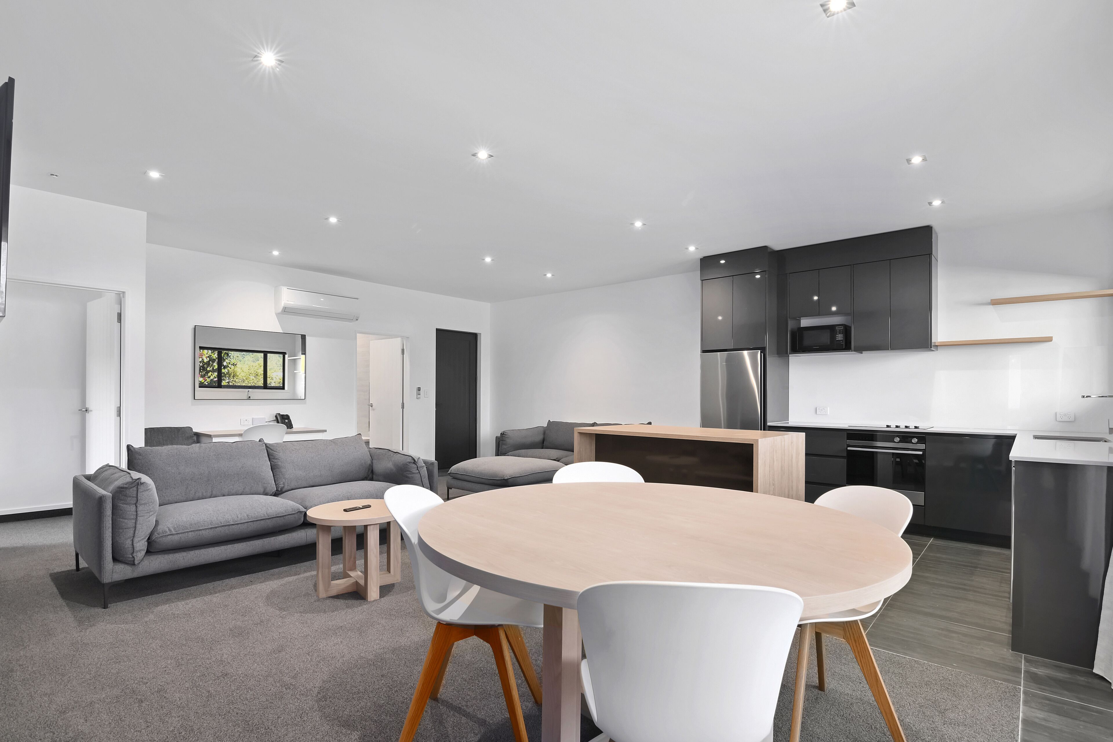 A modern living room with a grey sofa, a round wooden table, and white chairs. There is a kitchen area with dark cabinets and a microwave.