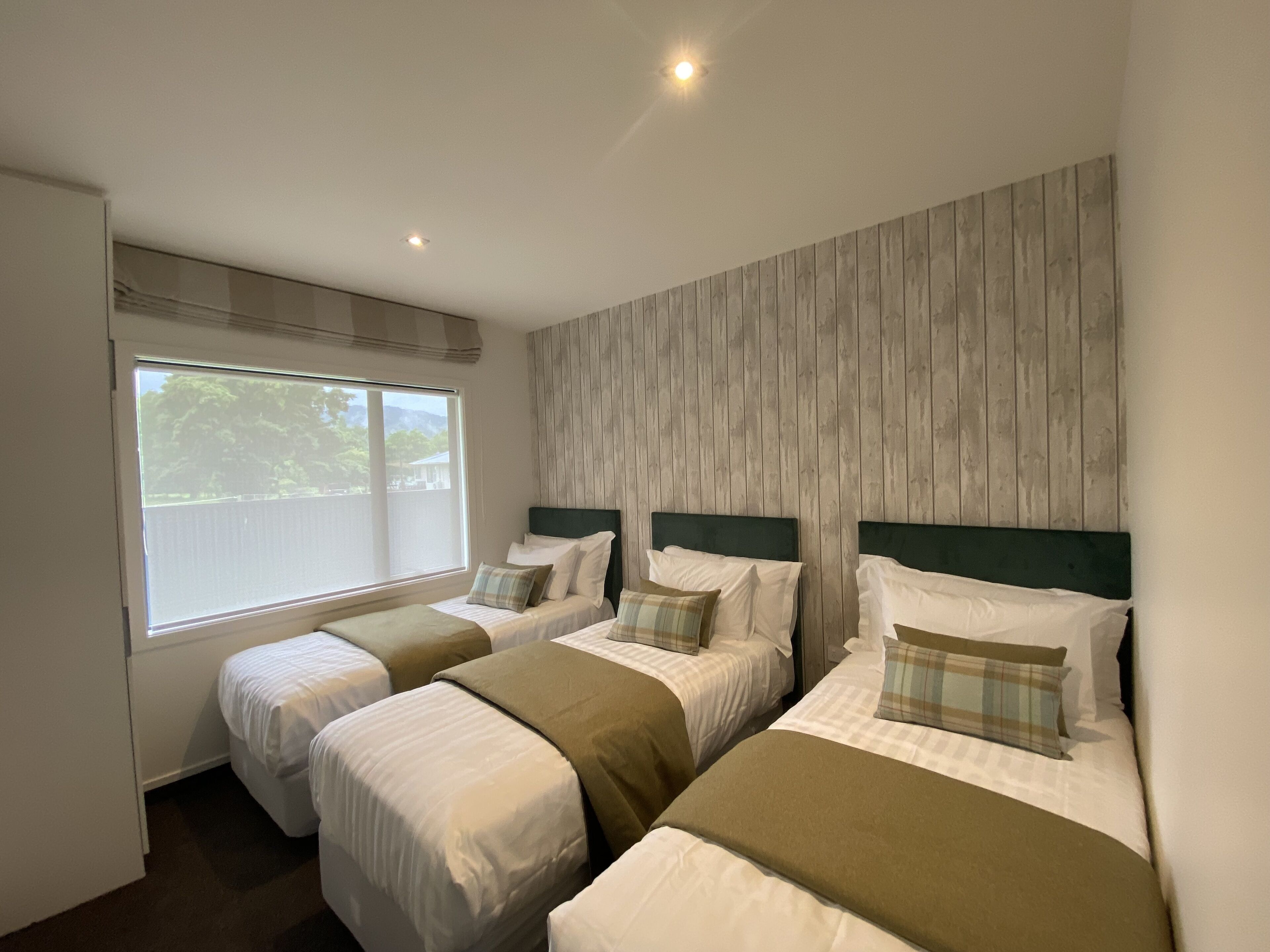 Habitación de hotel con tres camas individuales, un ventanal grande y un muro de paneles de madera.
