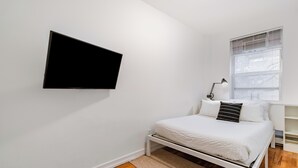 Ein Schlafzimmer mit Bett, Wandfernseher, Fenster mit Jalousien und Nachttisch mit Lampe.