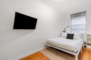 Quarto com cama, televisão na parede, janela com persianas e mesa de cabeceira com abajur.