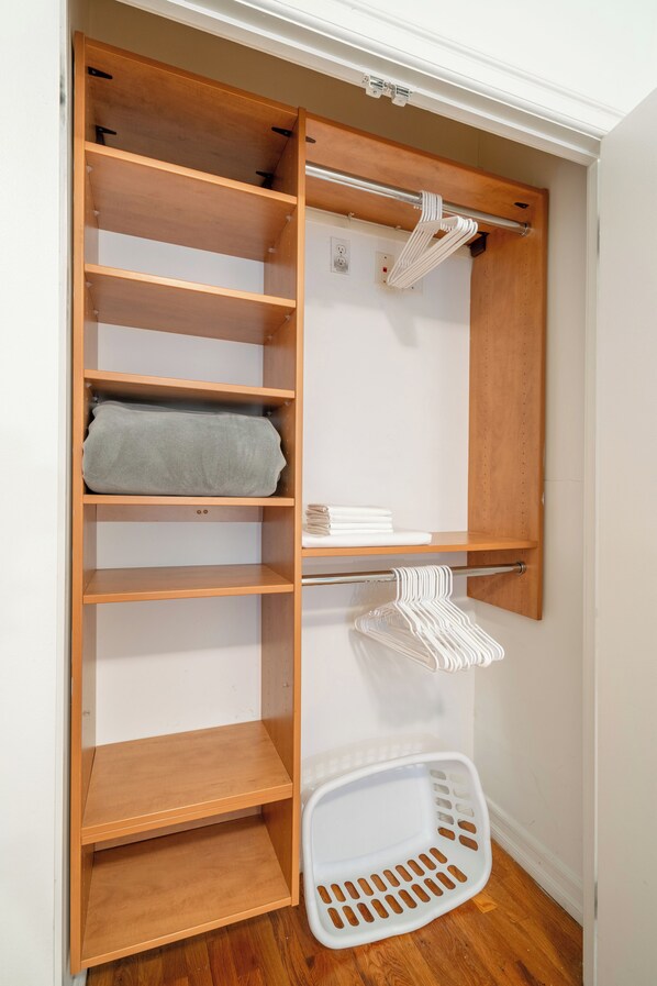 Un placard en bois avec plusieurs étagères et une tringle pour suspendre les vêtements, un panier à linge blanc et une serviette blanche posée sur l’étagère du haut.