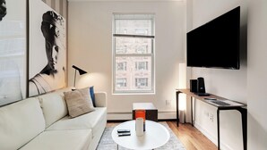 A modern living room with a white sofa, a coffee table, a flat-screen TV mounted on the wall, and a window with blinds.