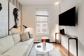Un salon moderne avec un canapé blanc, une table basse, un téléviseur à écran plat fixé au mur et une fenêtre avec des stores.