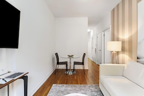 Uma sala de estar moderna com um sofá branco, uma mesa redonda com duas cadeiras, uma TV fixada na parede e um abajur sobre uma mesinha lateral.