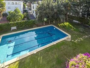 Ein rechteckiger Schwimmbereich mit Bahnen, umgeben von einer Rasenfläche mit Bäumen und einem weißen Gebäude im Hintergrund.