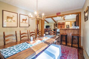 A dining area with a long wooden table, chairs, a chandelier, framed wildlife pictures, and a kitchen visible in the background.