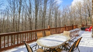 Ein mit Schnee bedeckter Deck mit einem runden Tisch und Stühlen, umgeben von einem Holgitter, mit Blick auf einen Wald.