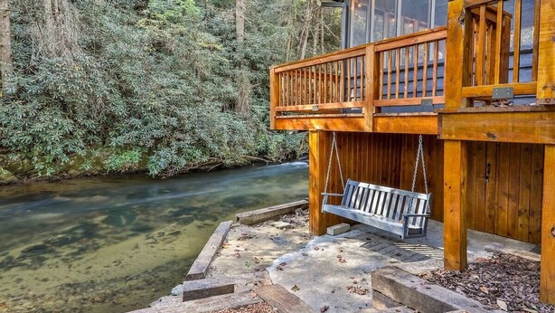 A wooden cabin with a balcony overlooking a river, surrounded by dense forest.