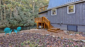A cabin with a wooden deck, a staircase, and two chairs by a stream.