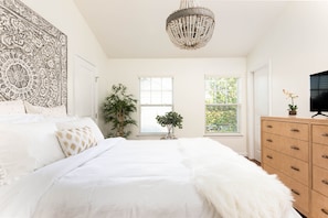 A bedroom with a large bed, a chandelier, a dresser, and a window with blinds.