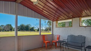 Eine überdachte Terrasse mit Holzboden, zwei orangefarbene Stühle, eine schwarze Metallbank und ein gemustertes Teppich.