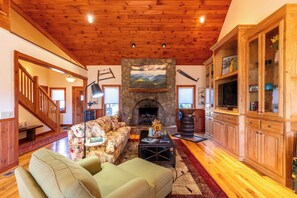 A living room with a stone fireplace, wooden ceiling, and a staircase. - A Tree Top Mountain Home - Views, Game Room, Pet Friendly (Banner Elk)