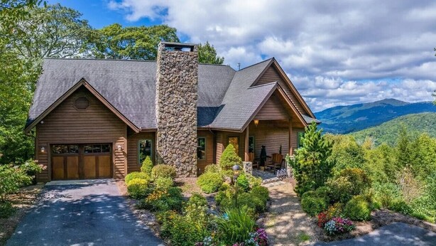 A two-story log cabin with a stone chimney, surrounded by lush greenery and mountains in the background.