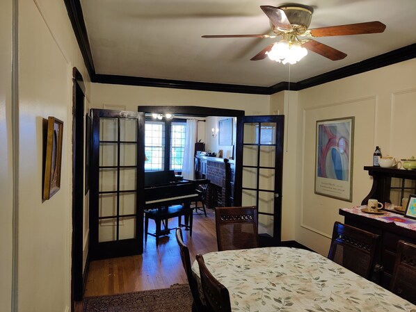 Una habitación con un piano, una mesa de comedor con sillas, un ventilador de techo y un cuadro en la pared.
