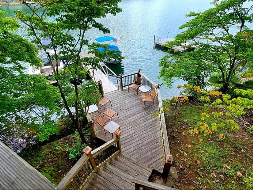 Un pontile in legno con mobili da esterno che si affaccia su un lago, barche ormeggiate e vegetazione lussureggiante.