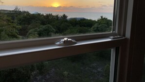 Blick aus dem Fenster auf einen Sonnenuntergang über einer Küstenlandschaft mit üppiger Vegetation und klarem Himmel.