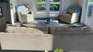 Ein geräumiges Wohnzimmer mit einer großen Couch, einem kleineren Sessel und einem Couchtisch. Es gibt einen Deckenventilator, ein großes Fenster mit Blick ins Freie und einen Blumentopf auf dem Boden.