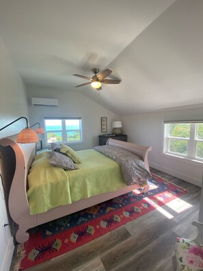 A bedroom with a bed, a ceiling fan, an air conditioning unit, a window, and a nightstand.