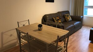 A living room with a wooden dining table and chairs, a brown leather sofa with patterned cushions, a black framed artwork on the wall, and a window with curtains. - Apex Wallace Street Apt With Free Parking (Glasgow)