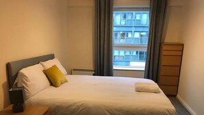 A neatly made bed with white linens and a single yellow pillow, a bedside table with a lamp, and a view of a building through a window with curtains. - Apex Wallace Street Apt With Free Parking (Glasgow)