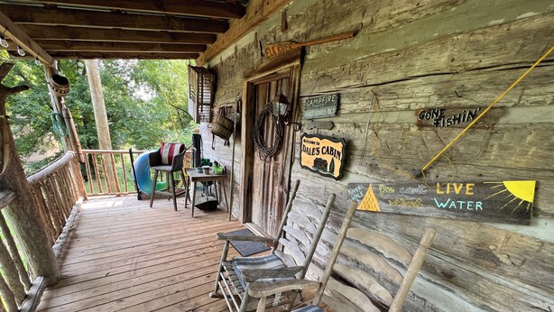 A rustic cabin with a wooden deck, a bench, and signs indicating a campfire and ghost stories. - Cheerful Riverfront Retreat (Fayetteville)