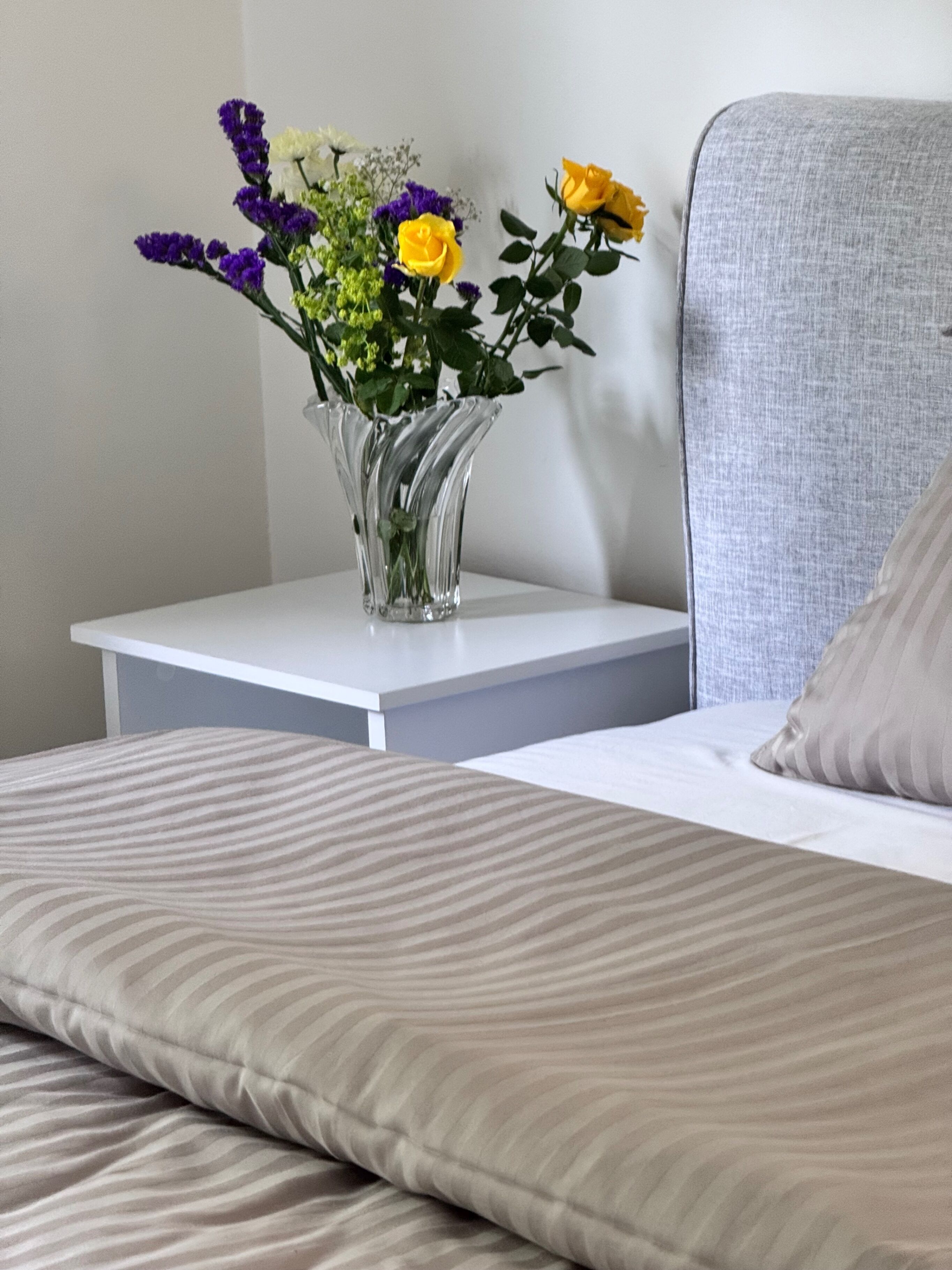 A bed with a striped pillow, a nightstand with a clear vase of flowers, and a grey upholstered headboard.