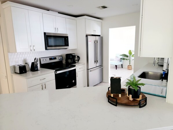 A modern kitchen with white cabinetry, a stainless steel refrigerator, a microwave, a stove, and a coffee maker. There is a small wooden table with potted plants.
