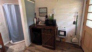 A small kitchen with wooden cabinets, a refrigerator, a microwave, and a coffee maker. There is a broom and a mop on the floor. - Nature's Tiny Riverside Hideaway (Fayetteville)