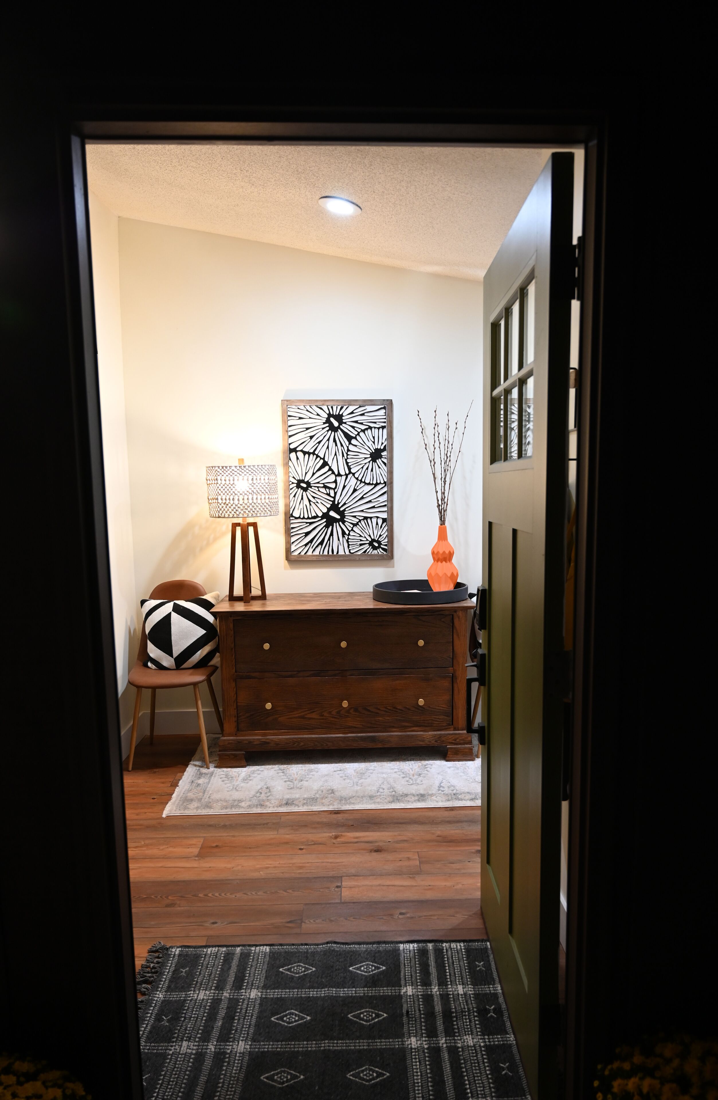 A hallway with a wooden floor, a patterned rug, a desk with a lamp, a decorative vase, and a wall art piece.
