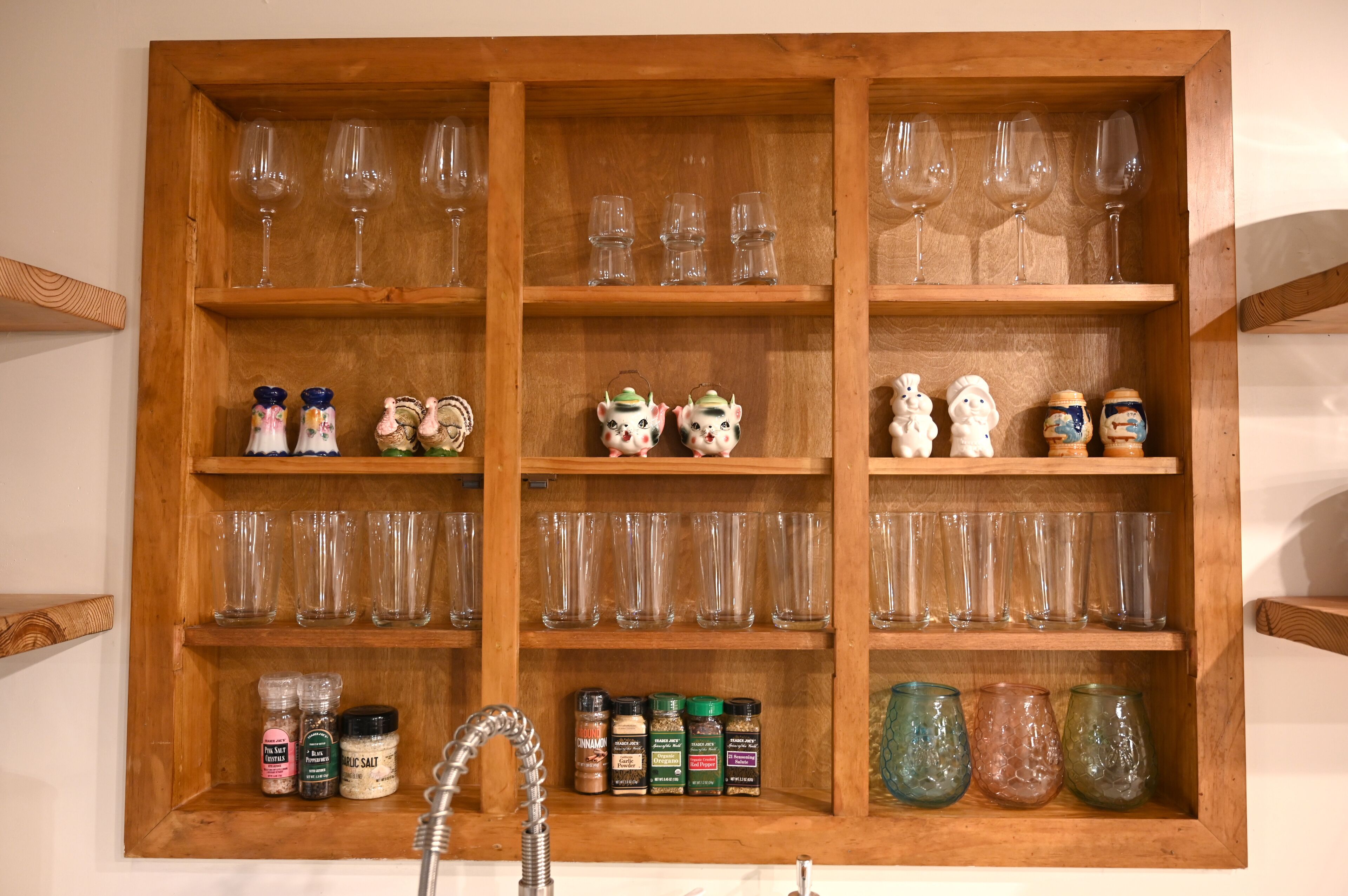 A wooden cabinet with multiple shelves containing wine glasses, decorative figurines, and spice jars.
