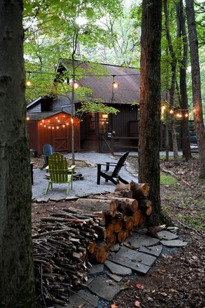A rustic cabin with a wooden deck, a stack of firewood, and string lights.