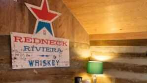 A rustic cabin interior with a wooden wall, a Redneck Riviera Whiskey sign, a small wooden bar, a heater, and a lamp. - Elk Riverside Escape (Fayetteville)