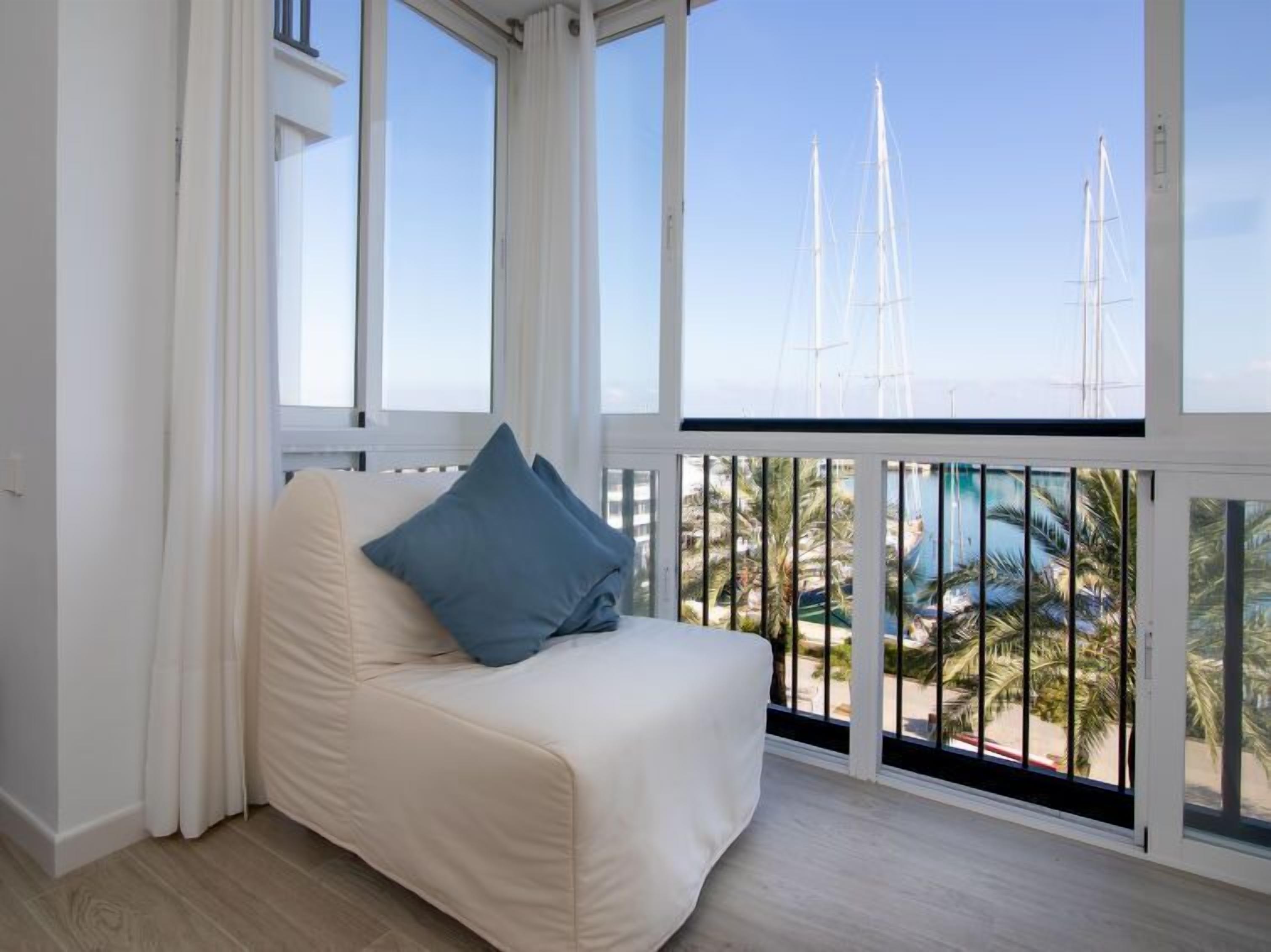 Ein Balkon mit einer weißen Couch und blauen Kissen mit Blick auf einen Yachthafen, wo Segelboote und Palmen zu sehen sind.