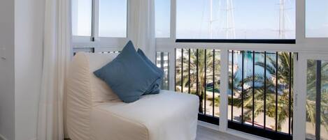 A balcony with a white sofa and blue pillows, overlooking a marina with sailboats and palm trees.