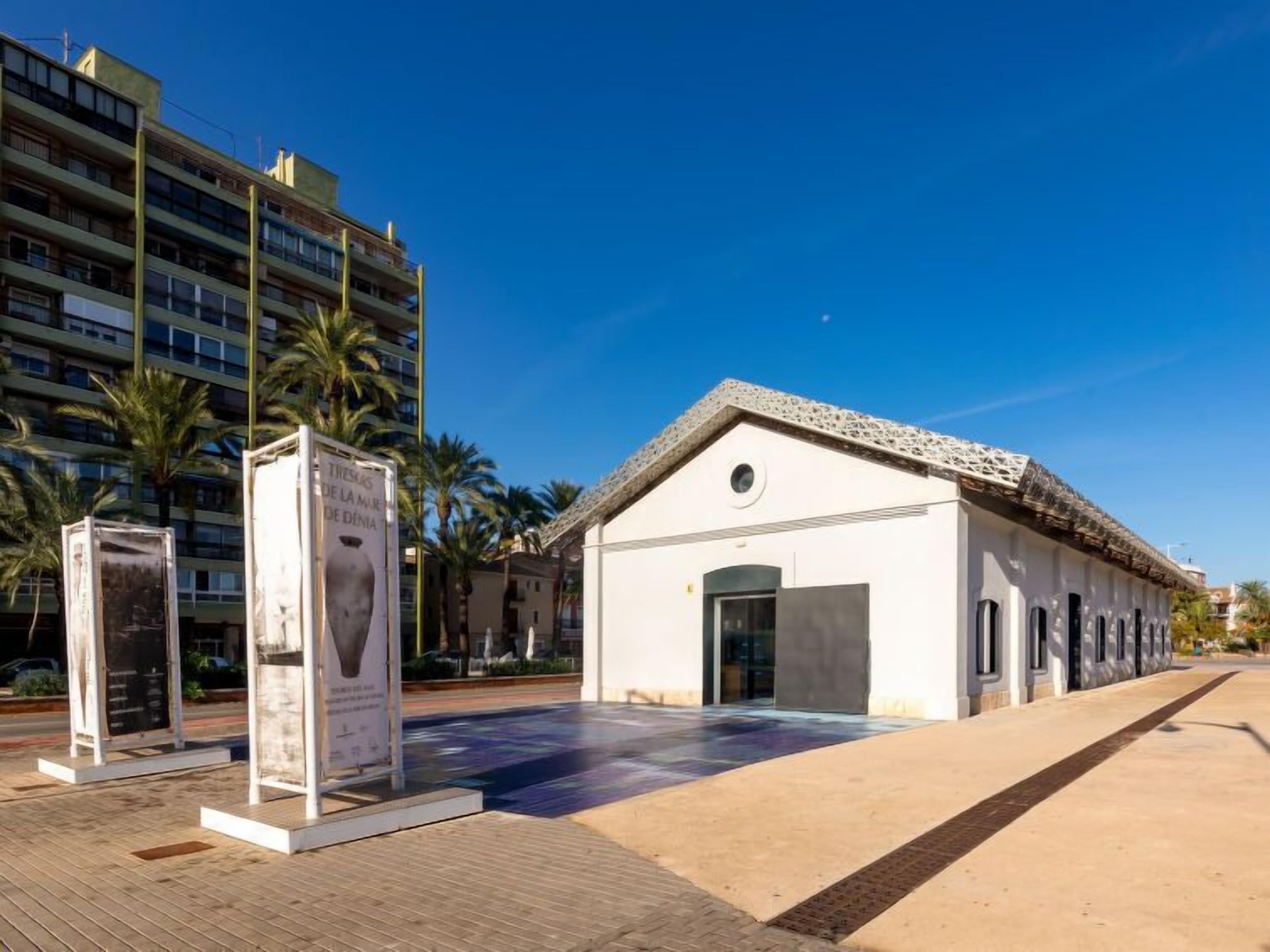 A building with a tiled roof and a small entrance. There are palm trees and a modern building in the background.