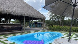 Ein Swimmingpool mit einer im Hintergrund sichtbaren, von einem Reetdach geschützten Struktur, umgeben von einer gefliesten Terrasse mit Grasflächen.