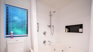 Une salle de bain équipée d’une baignoire, d’une douche et de toilettes. On y trouve une fenêtre avec des stores et une étagère contenant des articles de toilette.
